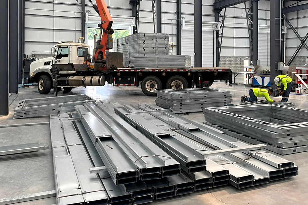 Zn-Al-Mg-coated steel C-profiles stacked on the plant floor, with a crane truck loaded with panels in the background